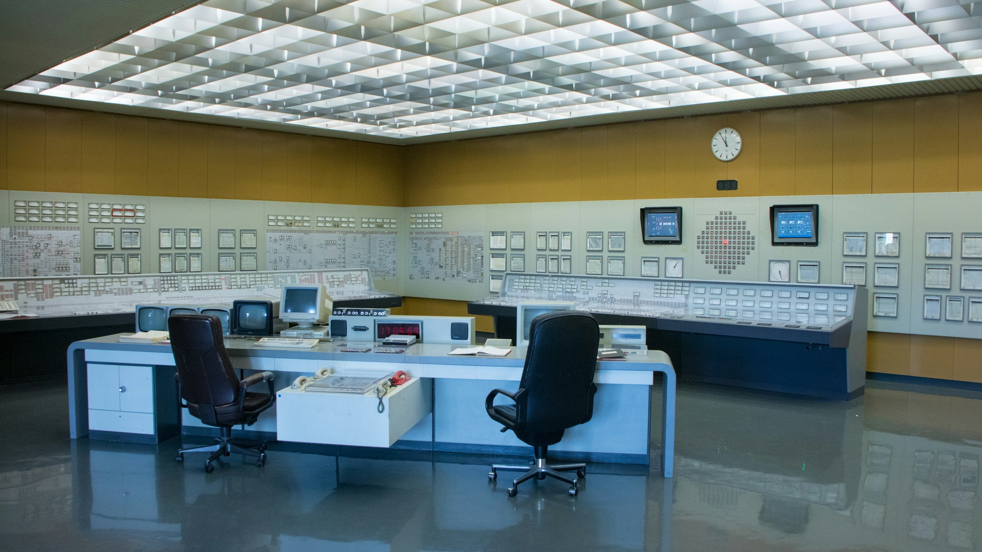 A plant control room: two operator chairs at a desk facing a wall of indicator panels and mimic diagrams.